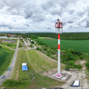Weitwinkelaufnahme eines hohen rot-weißen Mobilfunkmasts und einer künstlichen Baumspitze zur Tarnung. Im Hintergrund sind Felder, Wälder und ein kleines Dorf zu sehen.