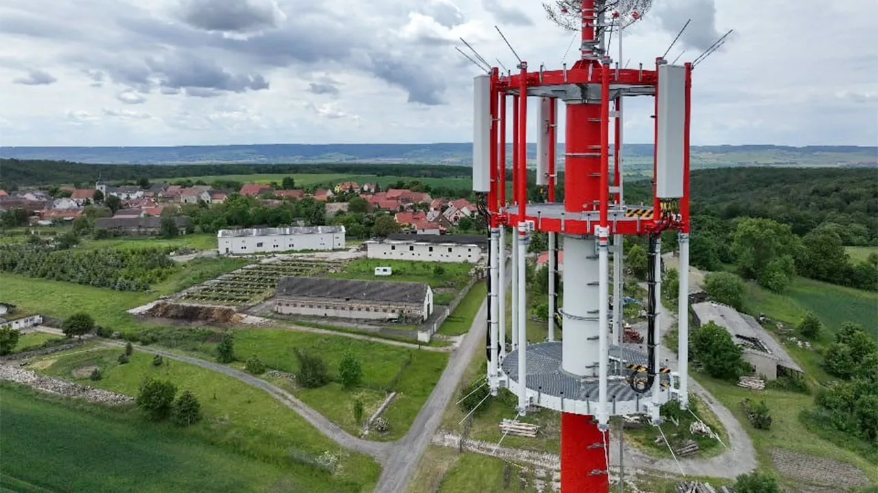 Nahaufnahme eines rot-weißen Mobilfunkmasts. Im Hintergrund sind Felder, Wälder und ein kleines Dorf zu sehen.